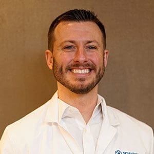 A man with short brown hair and a beard, smiling and wearing a white lab coat over a collared shirt, stands in front of a plain brown background.
