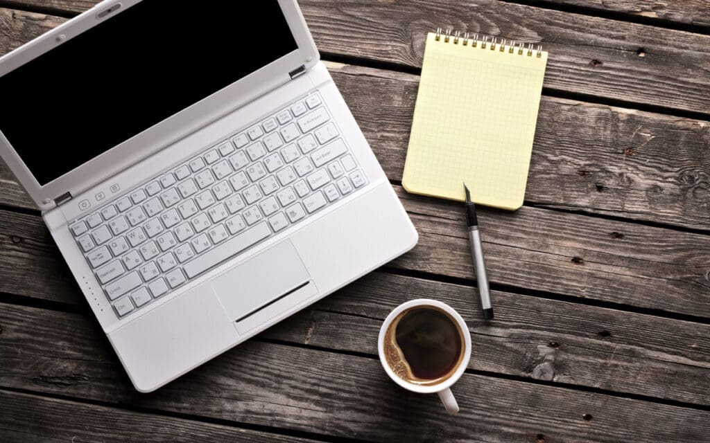A white laptop, a yellow notepad with a pen, and a cup of coffee sit on a rustic wooden table, ready for blog inspiration, viewed from above.