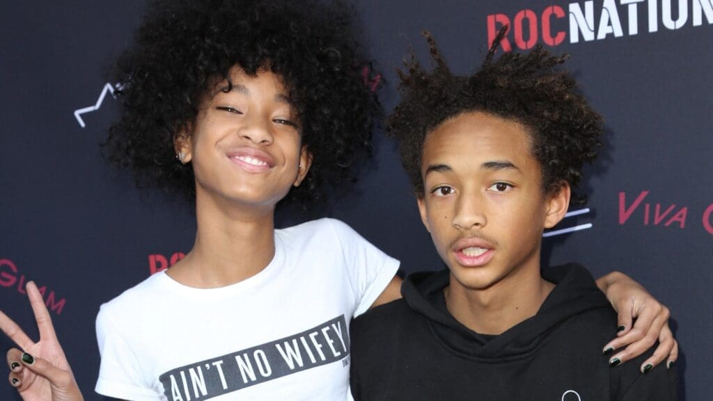 Two smiling Hollywood kids with natural curly hair pose on a red carpet. One, flashing braces and a peace sign in an "Aint No Wifey" tee, stands beside a friend in a black hoodie. The ROCNATION backdrop marks their rite of passage into the spotlight.
