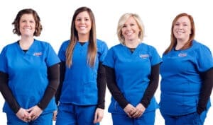 Four women wearing blue medical scrubs and black undershirts stand side by side, smiling at the camera, against a white background.