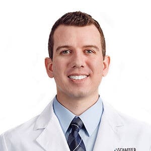 A man wearing a white lab coat, light blue dress shirt, and striped tie is smiling at the camera against a plain white background.
