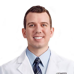 A man wearing a white lab coat, light blue dress shirt, and striped tie is smiling at the camera against a plain white background.