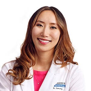 A woman with long wavy hair wearing a white lab coat and a pink top smiles at the camera against a white background.