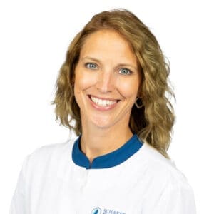 A woman with wavy light brown hair smiles at the camera. She is wearing a white coat with a blue collar and stands against a plain white background, representing a Lansing dentist.