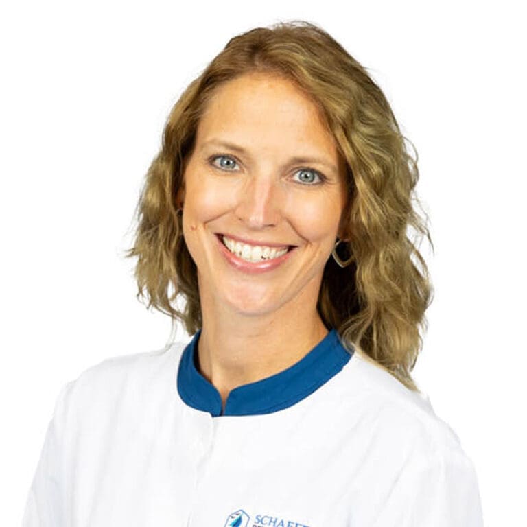 A woman with wavy light brown hair smiles at the camera. She is wearing a white coat with a blue collar and stands against a plain white background, representing a Lansing dentist.