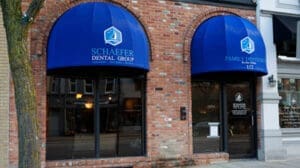 A brick building exterior with two large blue awnings that read Schaefer Dental Group and Family Dentistry above glass doors and windows. The street and nearby buildings are reflected in the glass.