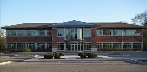 A two-story modern office building with large glass windows, a central entrance, and brick exterior, home to a dental clinic in Lansing, surrounded by a mostly empty parking lot and some small trees and shrubs.