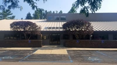 Single-story dental clinic with a metal roof and large front windows, flanked by two trees. Four accessible parking spaces are in front, and the sidewalk leads to a central glass entrance. Ideal for those seeking a Lansing dentist.