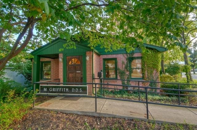 A small brick dental office with green trim, surrounded by trees and plants. A sign by the ramp entrance reads M. Griffith, D.D.S. Ivy climbs part of the building as sunlight filters through the leaves, highlighting this welcoming dentist in Lansing.