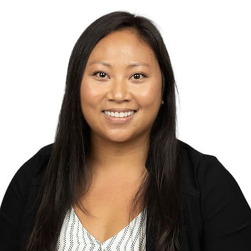 A woman with long dark hair smiles at the camera. She is wearing a black blazer over a patterned top, and the plain white background gives this Lansing dentist a professional and approachable look.