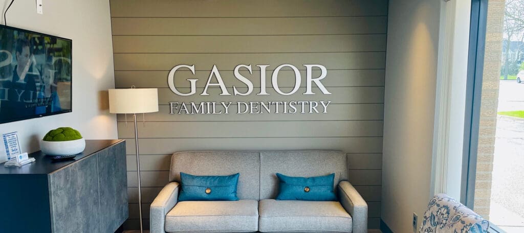 A waiting area at a dental clinic with a gray sofa, two teal pillows, a floor lamp, a cabinet with a bowl of green decorative spheres, and a large sign on the wall that says GASIOR FAMILY DENTISTRY.