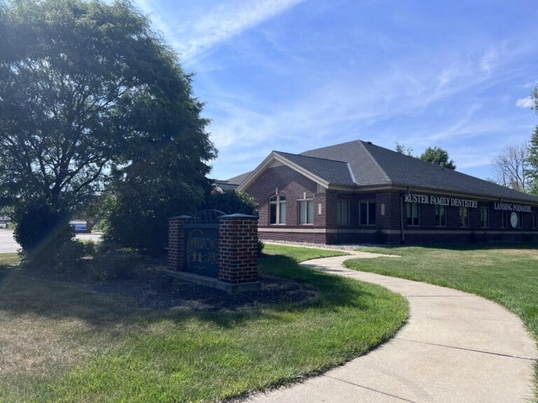 A brick building with signs for Foster Family Dentistry and Lansing Pediatric is surrounded by grass, a tree, and a sidewalk on a sunny day. A brick sign labeled Merrion Professional stands near the path, highlighting this dentist in Lansing.