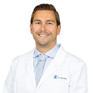 A smiling man with short brown hair wears a white lab coat reading Dr. Gosselin, a light blue shirt, and striped tie. The blue logo hints he’s a dentist in Lansing. The background is plain white.