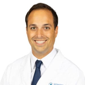 A man with short brown hair, wearing a white medical coat, white shirt, and dark blue tie, smiles at the camera against a plain white background—perfectly representing a friendly Lansing dentist.
