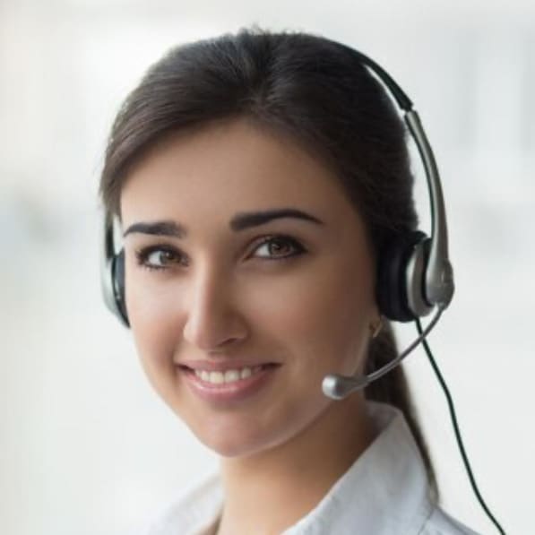A woman with long brown hair, wearing a headset with a microphone, smiles at the camera. She is dressed in a white collared shirt and appears to be in a bright, professional dental clinic.