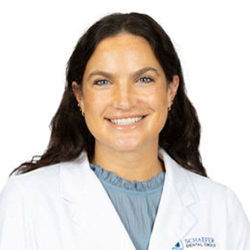 Smiling woman with long brown hair wearing a white lab coat and blue blouse, posing against a plain white background, representing a friendly Lansing dentist.