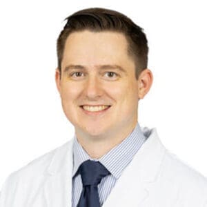A smiling man with short brown hair wearing a white lab coat, blue tie, and light blue striped shirt stands against a plain white background, representing a friendly dentist in Lansing.