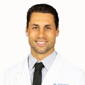 A smiling man with short dark hair, wearing a white lab coat, patterned shirt, and black tie, poses in front of a plain white background, representing a friendly Lansing dentist.