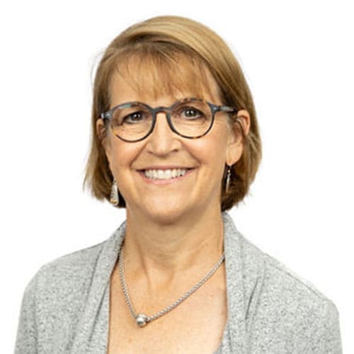 A smiling middle-aged woman with short light brown hair, wearing round eyeglasses, silver earrings, a necklace, and a light gray sweater, stands against a plain white background—an approachable Lansing dentist.