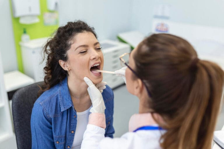 A dentist in Lansing examines a woman's throat using a tongue depressor in a dental clinic. The woman sits with her mouth open while the professional, wearing gloves and a white coat, looks into her mouth.