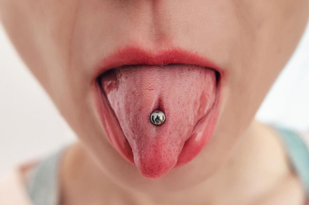 A close-up of a person's mouth with their tongue sticking out, displaying a silver ball tongue piercing. The lips are slightly parted, and the background is out of focus—a unique image often seen by a dentist in Lansing.