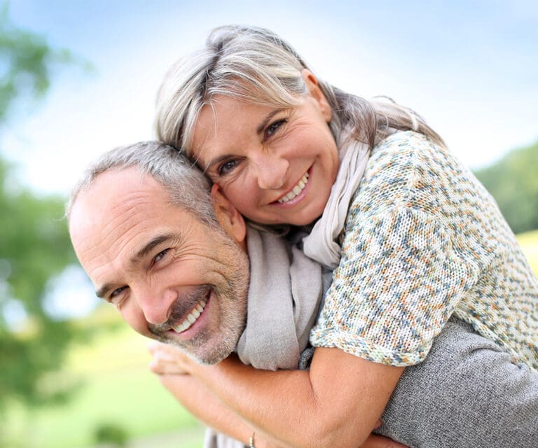 A smiling middle-aged woman with gray hair embraces a joyful man from behind outdoors on a sunny day, with a blurred green and blue background, highlighting the happy couple after their visit to the Lansing dentist.