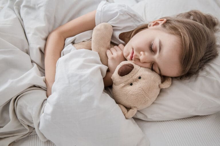 A young girl with light brown hair sleeps peacefully in bed, hugging a beige teddy bear and wrapped in white blankets, dreaming of her next visit to the caring Lansing dentist.