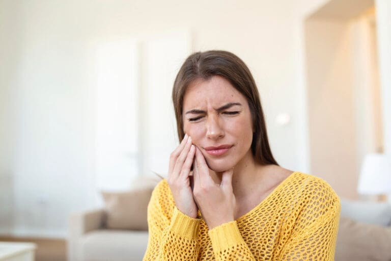 A young woman in a yellow sweater sits indoors, holding her cheek and closing her eyes in discomfort, likely needing help from a Lansing dentist for tooth or jaw pain.