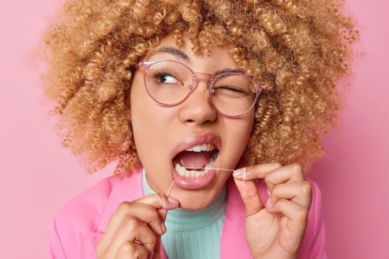 A woman with curly blonde hair and glasses flosses her teeth while winking, wearing a pink blazer and turquoise top, against a pink background, promoting good habits encouraged by your local Lansing dentist.