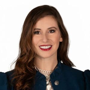 Dr Heather Rae McAlvey, a woman with long, wavy brown hair, smiles at the camera. She is wearing a navy jacket over a white lace top, set against a white background.