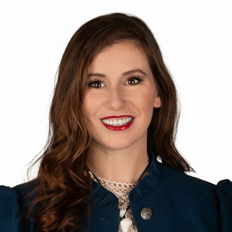 Dr Heather Rae McAlvey, a woman with long, wavy brown hair, smiles at the camera. She is wearing a navy jacket over a white lace top, set against a white background.