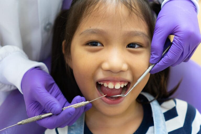 A young girl sits in a dental chair, smiling with her mouth open as a Pediatric Dentist in Lansing MI, wearing purple gloves, examines her teeth with dental tools.