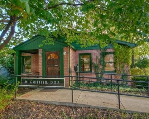 A small brick dental office with green trim, surrounded by trees and plants. A sign by the ramp entrance reads M. Griffith, D.D.S. Ivy climbs part of the building as sunlight filters through the leaves, highlighting this welcoming dentist in Lansing.