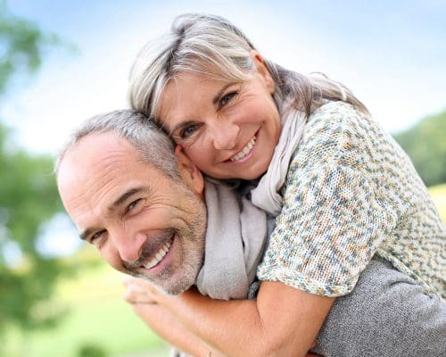 A smiling middle-aged woman with gray hair embraces a joyful man from behind outdoors on a sunny day, with a blurred green and blue background, highlighting the happy couple after their visit to the Lansing dentist.