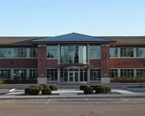 A two-story modern office building with large glass windows, a central entrance, and brick exterior, home to a dental clinic in Lansing, surrounded by a mostly empty parking lot and some small trees and shrubs.