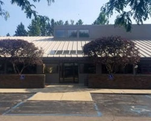 Single-story dental clinic with a metal roof and large front windows, flanked by two trees. Four accessible parking spaces are in front, and the sidewalk leads to a central glass entrance. Ideal for those seeking a Lansing dentist.