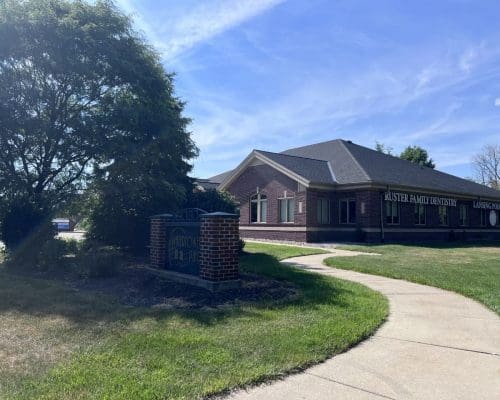A brick building with signs for Foster Family Dentistry and Lansing Pediatric is surrounded by grass, a tree, and a sidewalk on a sunny day. A brick sign labeled Merrion Professional stands near the path, highlighting this dentist in Lansing.