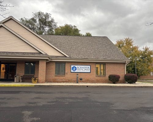 A one-story brick dental clinic with a sign reading Schaefer Dental Group stands near shrubs by the entrance under cloudy skies. The parking lot is empty.
