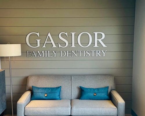 A waiting area at a dental clinic with a gray sofa, two teal pillows, a floor lamp, a cabinet with a bowl of green decorative spheres, and a large sign on the wall that says GASIOR FAMILY DENTISTRY.